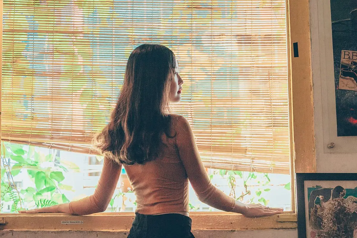 Một người đứng bên cửa sổ có rèm tre, đang suy nghĩ