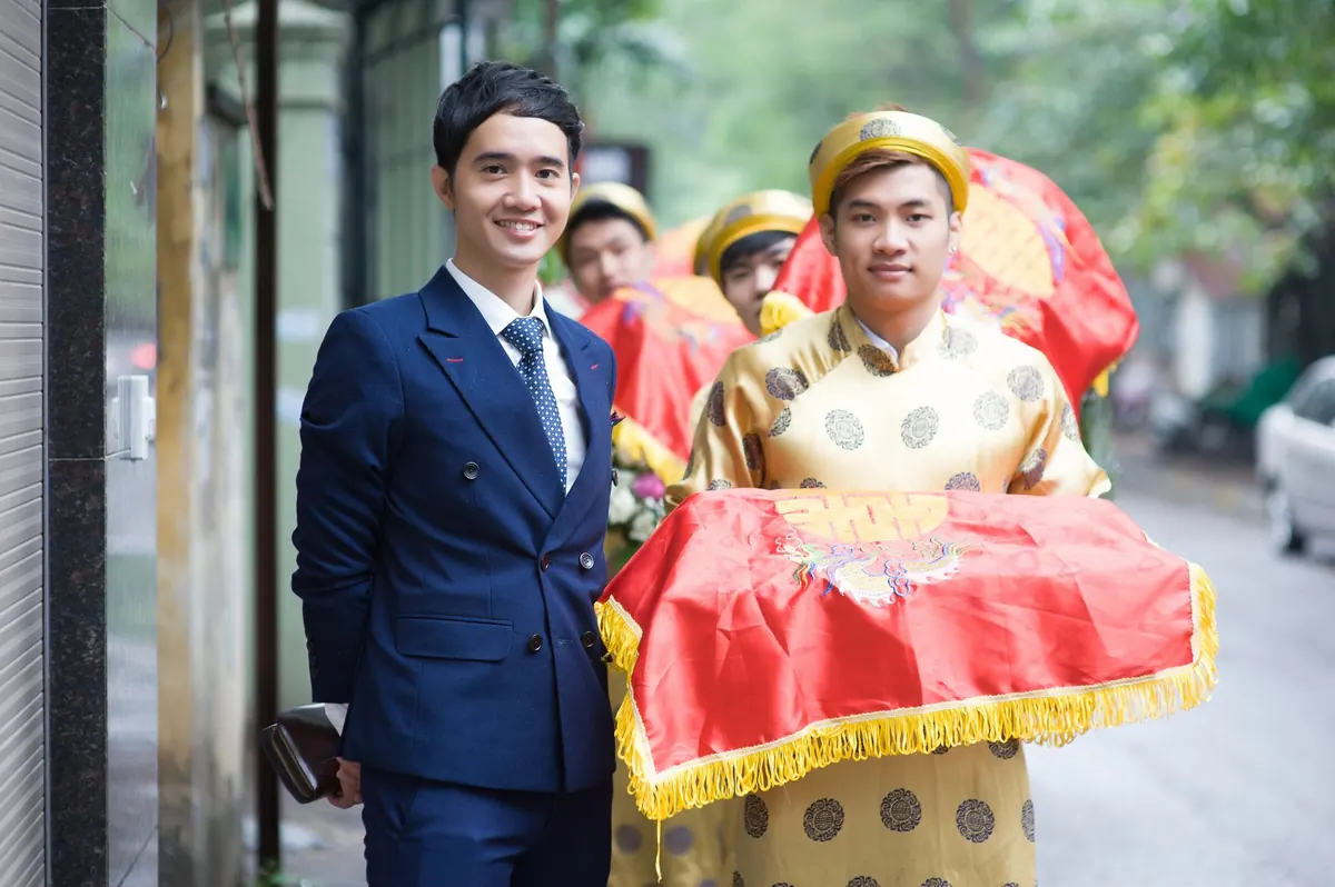 Chú rể mặc vest đứng cạnh đội bê tráp áo the vàng trên con ngõ