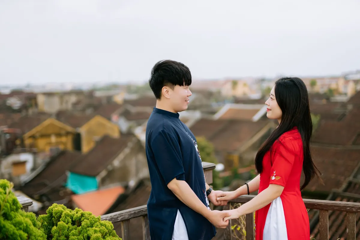 Đôi bạn trẻ mặc áo dài nắm tay nhau trên ban công nhìn xuống mái ngói phố cổ