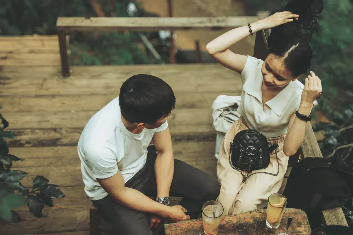 Một đôi ngồi trên bậc gỗ, người con gái vuốt tóc, hai ly nước để trên bàn