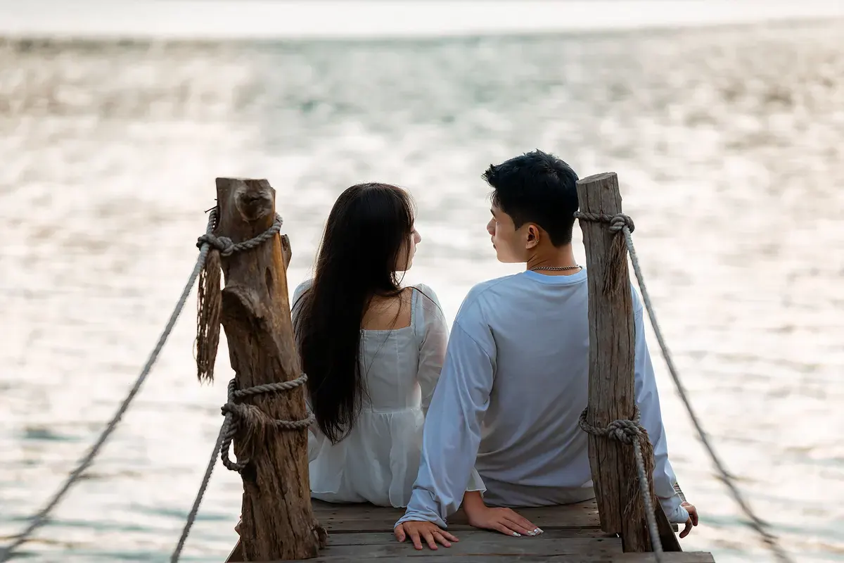 Một đôi ngồi cạnh nhau trên cầu gỗ bên mặt nước, nhìn về phía nhau