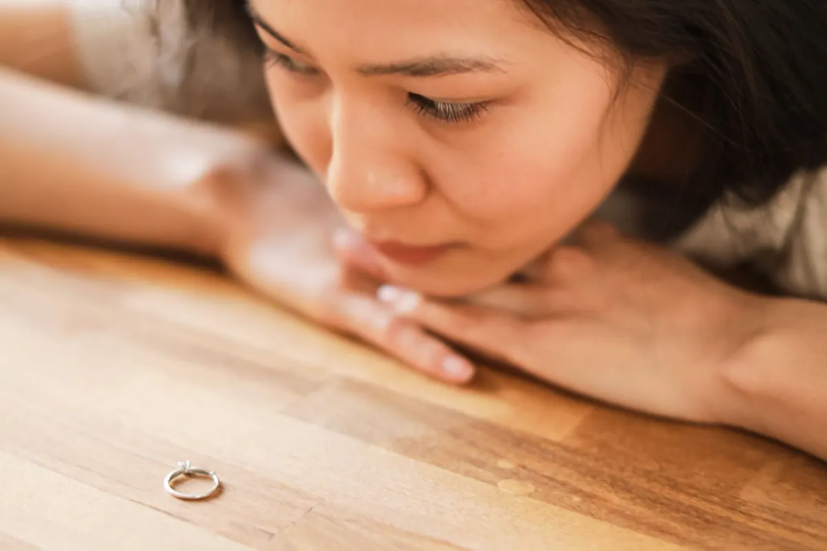 Người phụ nữ tựa cằm lên bàn gỗ, trầm ngâm nhìn chiếc nhẫn cưới trước mặt