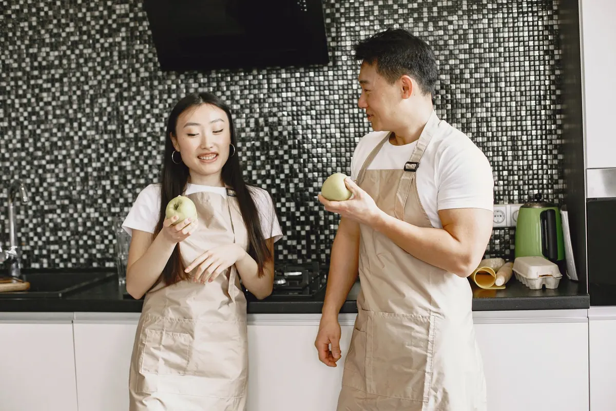 Một cặp đôi đeo tạp dề đứng trò chuyện trong bếp, mỗi người cầm một quả táo xanh