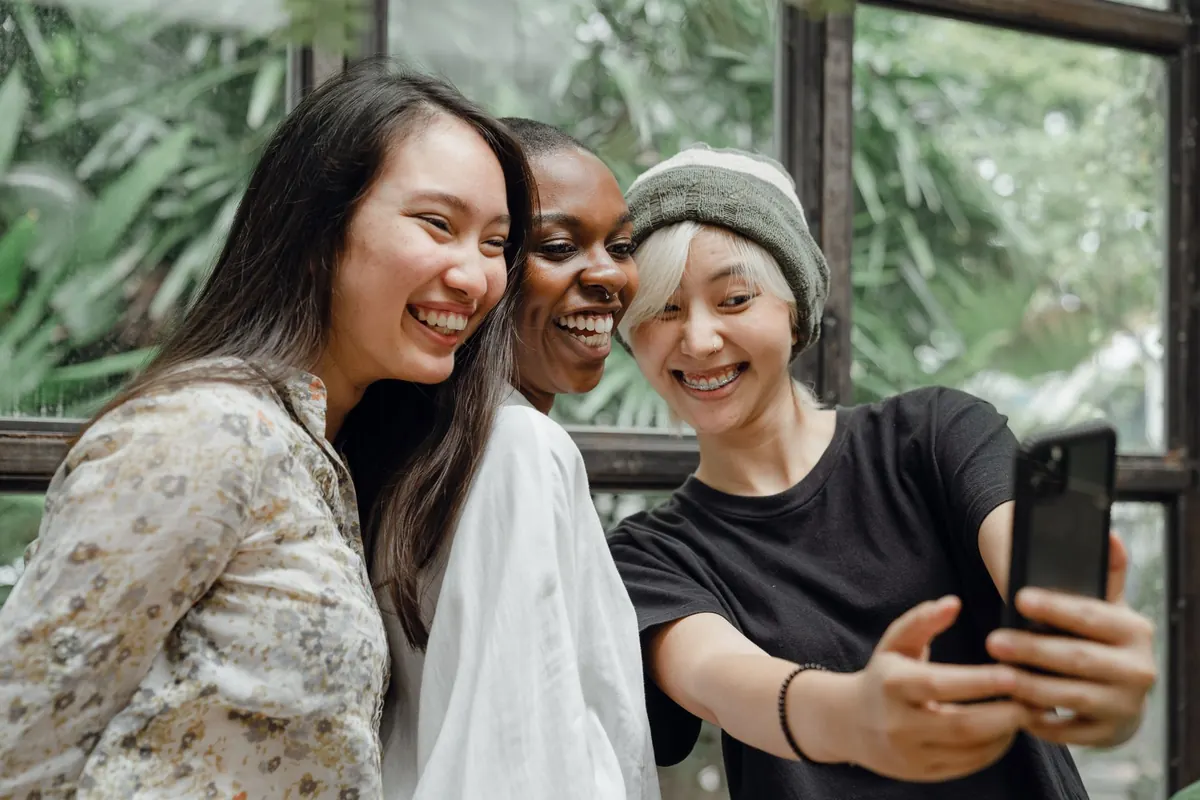 Ba cô gái cười tươi chụp ảnh selfie bằng điện thoại cạnh ô cửa kính nhiều cây xanh