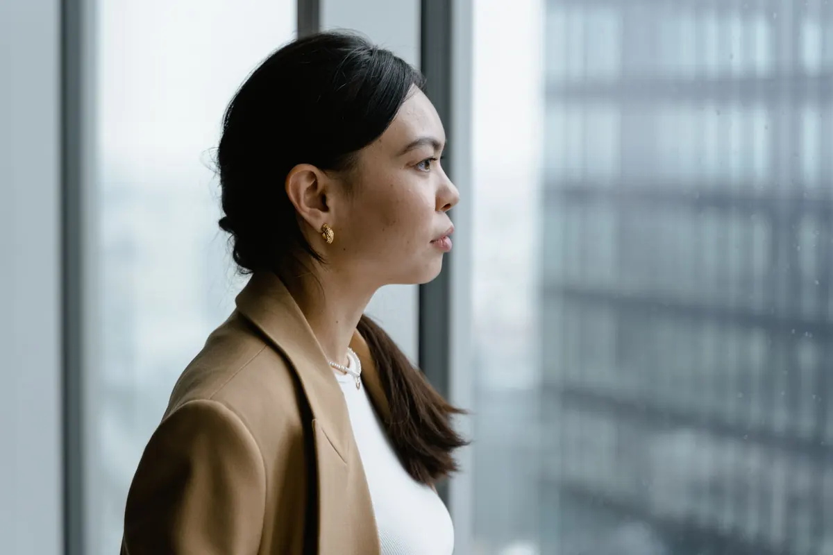 Người phụ nữ mặc áo vest nâu đứng bên cửa kính tòa nhà, nhìn ra ngoài suy nghĩ
